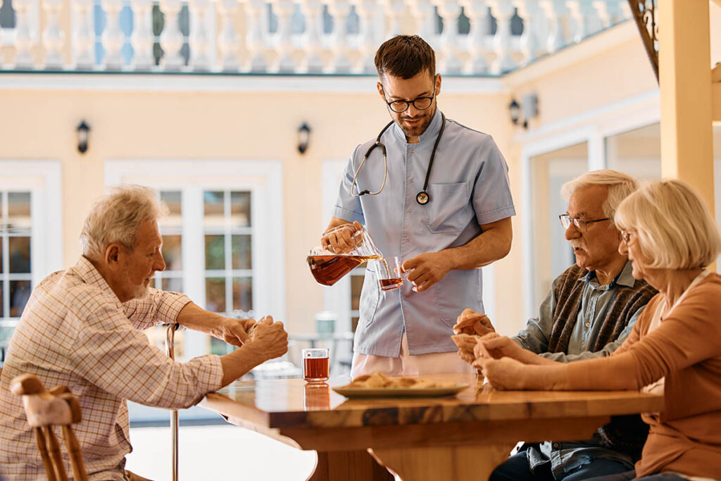 Novellus Kingwood | Male caregiver pouring drinks to group of seniors who are playing cards at nursing home.