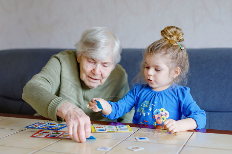 Novellus Kingwood | Beautiful toddler girl and grand grandmother playing together pictures lotto table cards game at home. Cute child and senior woman having fun together. Happy family indoors