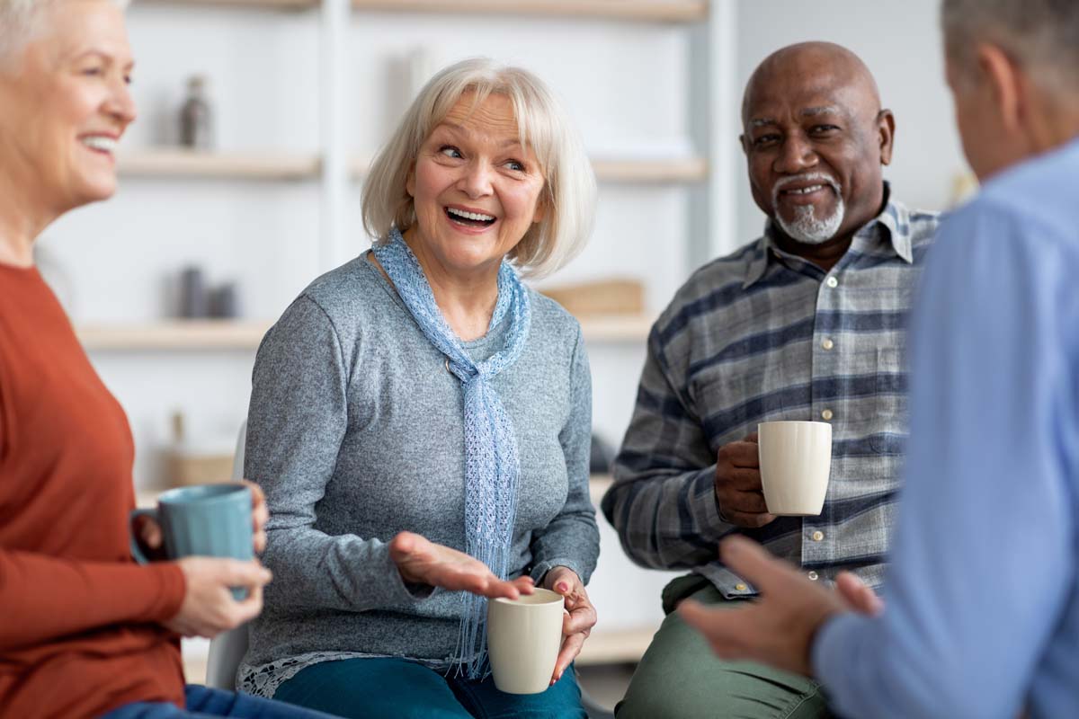 Orangeburg Memory Care | Emotional seniors watching television in the living room Group of positive seniors drinking tea and chatting