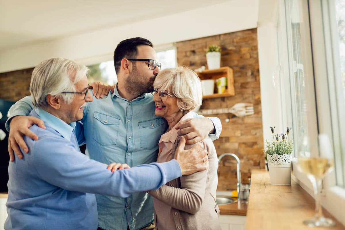 Camlu Assisted Living | Adult son and his senior parents hugging and smiling in the kitchen